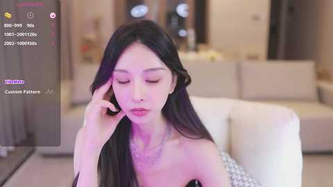 Media: Video of a young East Asian woman with long black hair, wearing a black strapless dress, sitting on a white sofa in a modern living room.