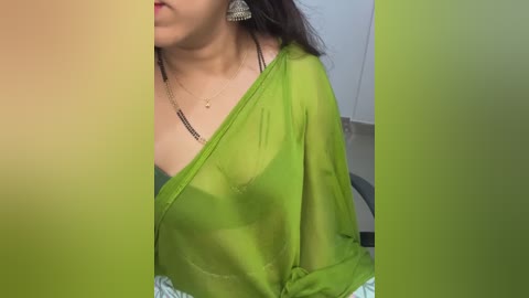 Media: A video of a woman with medium skin tone and long dark hair, wearing a sheer, green, one-shouldered top, revealing a black bra underneath. She has dangling earrings and sits indoors, partially blurred background.