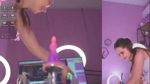 Media: Video of a young woman with light skin and dark hair in a pink room, using a pink dildo and looking at a computer screen, surrounded by glowing pink lights and hexagonal wall art.