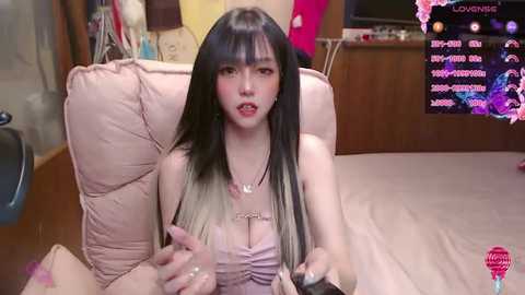 Media: Video of an East Asian woman with long, black-and-blonde hair, wearing a pink, sleeveless top, seated on a beige couch, holding a video game controller, in a cozy, cluttered living room.