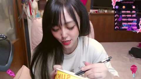 Media: Video of an East Asian woman with long black hair, wearing a white shirt, writing in a notebook. Background includes a wooden desk, monitor displaying text, and a pink bag.