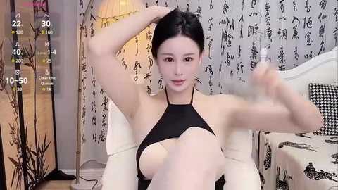 Media: A video of an East Asian woman with fair skin and black hair, wearing a black halter top, sitting on a bed with white bedding and black patterned pillows. She holds a water bottle, in a cozy, dimly-lit room with a lamp.