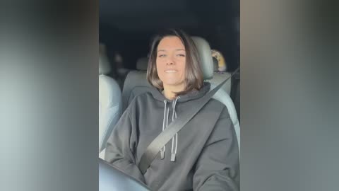 Media: Video of a young woman with shoulder-length brown hair, wearing a dark hoodie, seated in a car, with a seatbelt across her chest.