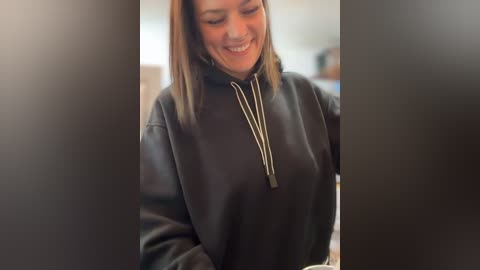 Media: Video of a smiling, light-skinned woman with shoulder-length straight brown hair, wearing a black hoodie with a long chain necklace. Background is blurred, indoors.