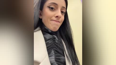 Media: Video of a Latina woman with long, straight black hair, wearing a white puffy jacket, black ruffled top, and black eyeliner. She smiles slightly, set against a blurred background of light colors.