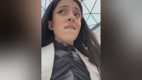 Media: A video of a young woman with long black hair, wearing a puffy black jacket, standing in a modern, glass-walled room with triangular windows. Her expression is neutral, and her makeup includes winged eyeliner.