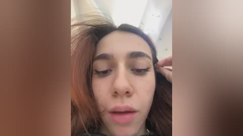 Media: Video of a young woman with medium brown skin, long dark hair, and full eyebrows, applying eyeliner in a bathroom with visible ceiling lights and blurred background.