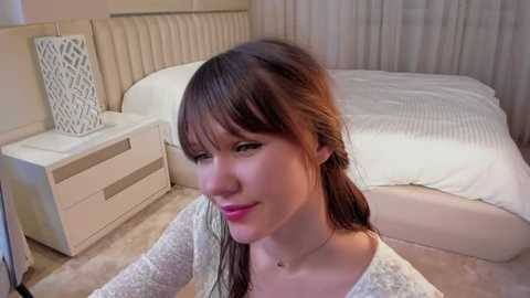 Media: Video of a smiling, fair-skinned woman with long brown hair, wearing a white sweater, sitting on a beige carpet near a white bed with a patterned lamp on a bedside table.