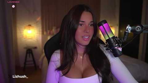 Media: Video of a woman with long, dark hair and large breasts, wearing a white top, singing into a microphone. The background shows a dimly lit bedroom with a nightstand lamp.