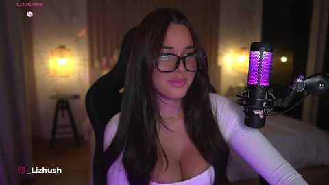 Media: Video of a fair-skinned woman with long dark hair and large glasses, wearing a white top, seated in a black chair, recording herself with a purple light on a microphone.