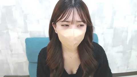 Media: Video of a young East Asian woman with long brown hair, wearing a black sweater, a surgical mask, and sitting in a blue chair. The background features a blurred, light-colored curtain.