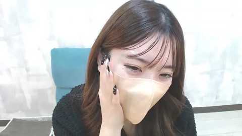 Media: Video of an Asian woman with long brown hair, wearing a surgical mask, black earrings, and a black sweater, seated in a room with white curtains and a blue chair.