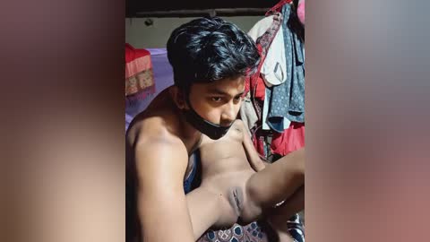 Media: Video of a shirtless South Asian man with a bound mouth, performing oral sex on a woman. Background features a cluttered room with clothes hanging.