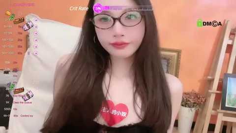 Media: Video of a slender, fair-skinned woman with long, straight brown hair, wearing glasses and a black top, holding a red heart-shaped sign. Background features an orange wall, a white chair, and a wooden shelf.