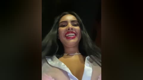 Media: Video of a smiling woman with long dark hair, wearing a white shirt, black choker, and red lipstick, set against a dim background.