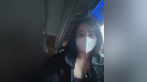 Media: A blurry video of a woman in a black leather jacket, wearing a white face mask, seated inside a dimly lit vehicle.