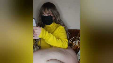 Media: A video of a woman with long, wavy brown hair, wearing a yellow turtleneck, black face mask, and holding a phone, sitting on a patterned couch.