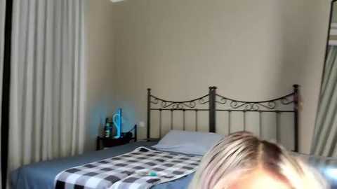 Media: A video of a bedroom featuring a bed with a black wrought-iron headboard, white pillows, a gray and white checkered blanket, and a blue statue on a nightstand. The room has beige walls and sheer white curtains.