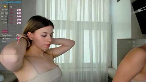 Media: A video of a young, fair-skinned woman with straight brown hair, wearing a beige strapless dress, adjusting her hair in a modern, minimalist room with white curtains and a flat-screen TV.