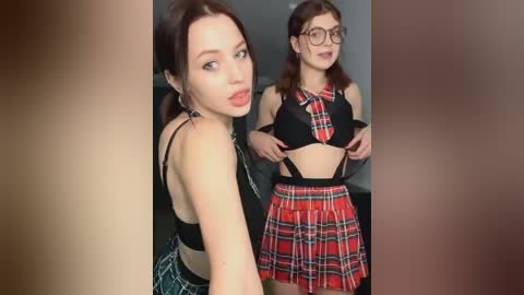 Media: Video of two women in matching red plaid outfits: one in a black bra, the other in a black top and pleated skirt. Both have fair skin, brunette hair, and glasses. Background is blurred.