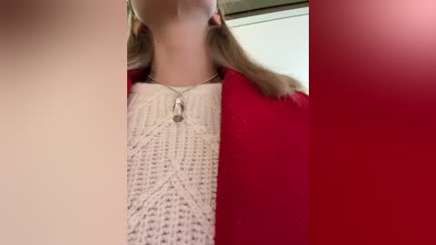 Media: A video of a woman with fair skin, wearing a white knit sweater and red cardigan, her blonde hair in pigtails, standing against a white wall.