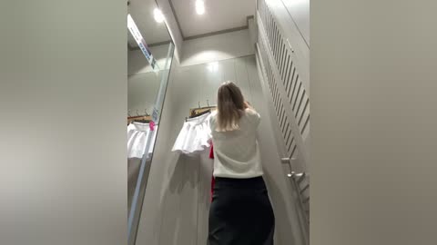 Media: A video captures a woman in a dressing room, facing away, with long blonde hair. She wears a white sweater and black pants, holding a white shirt. The room has mirrored walls and overhead lighting, displaying other clothing items.