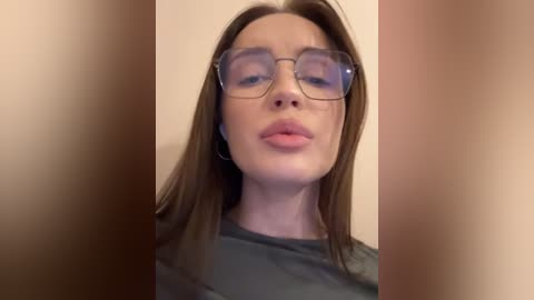 Media: Video of a fair-skinned woman with straight, shoulder-length brown hair, wearing clear-rimmed glasses and a grey top, against a blurred, beige background.