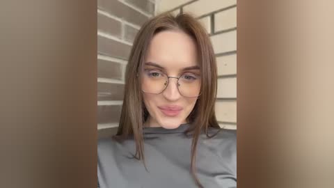 Media: Video of a young woman with fair skin, brown hair, and glasses, wearing a grey turtleneck sweater. She stands in front of a beige brick wall, smiling slightly.