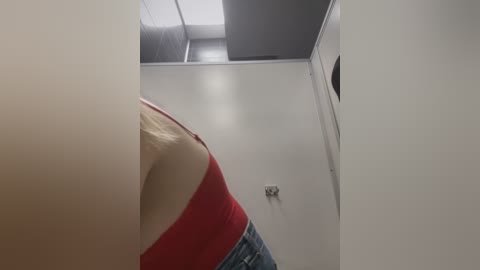 Media: Video of a woman's upper body in a dimly lit bathroom. She has fair skin and wears a red tank top, blue jeans, and is seen from the side.