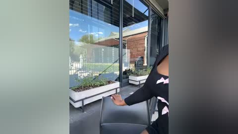 Media: Video of a person with medium skin tone, wearing a black off-shoulder top, touching a windowpane with their left hand, inside a modern, glass-walled room with potted plants, a brick building, and a clear blue sky visible outside.