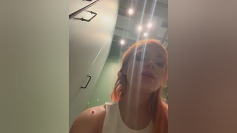 Media: Video of a fair-skinned woman with shoulder-length, light orange hair, wearing a white tank top, standing in a modern bathroom with a glass shower door.