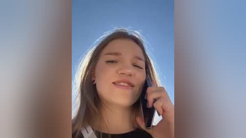 Media: Video of a young Caucasian woman with light skin and long, straight brown hair, smiling while holding a phone to her ear against a clear blue sky background.