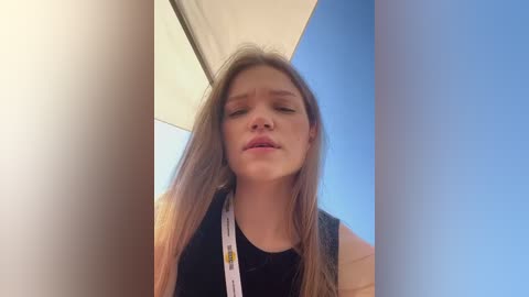 Media: Video of a young woman with long, light brown hair and pale skin, wearing a black top and lanyard, standing in a bright, blue sky setting.