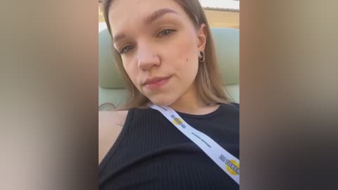 Media: Video of a young woman with light skin and brown hair, wearing a black ribbed tank top, seated in a car with a seatbelt across her chest.