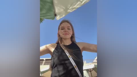 Media: Video of a young Caucasian woman with light skin and long brown hair, wearing a black tank top, standing outdoors under a bright blue sky with a green and white umbrella.