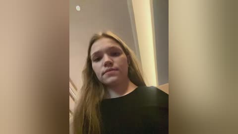 Media: A video of a young woman with long, straight, light brown hair, wearing a black shirt, standing in a dimly lit hallway with beige walls and a recessed ceiling light.