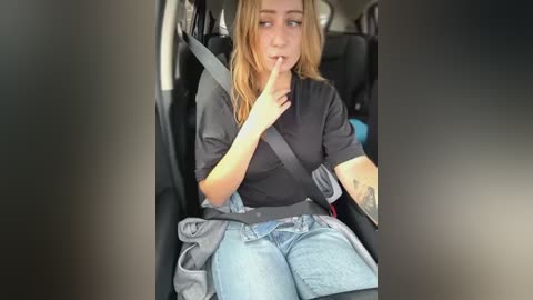 Media: Video of a young woman with long blonde hair, wearing a black top and blue jeans, sitting in a car with a seatbelt, holding her finger to her lips.