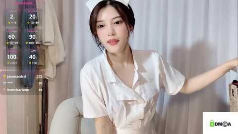 Media: Video of an East Asian woman with light skin and brown hair, wearing a white nurse uniform and cap, standing in a hospital room with a white curtain backdrop.