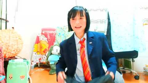 Media: A video of a young Asian man with black hair, wearing a blue suit, red tie, and a blue shirt, smiling in a cluttered, brightly lit room with toys, stuffed animals, and a chair.