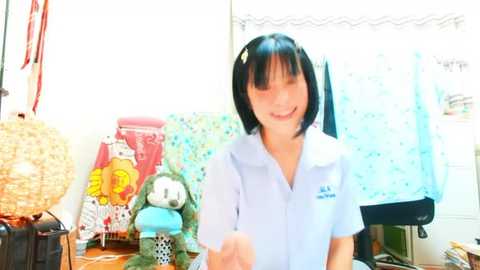 Media: A video of a young Asian girl with short black hair, wearing a light blue uniform shirt, smiling and holding a drink, surrounded by a cluttered room with toys, a wicker lamp, and colorful fabric.