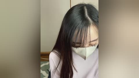 Media: Video of an Asian woman with long black hair, wearing a white mask and a light-colored sweater, sitting with her head down. The background features neutral-toned walls and a wooden door.