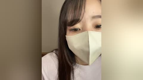 Media: Video of an Asian woman with long black hair, wearing a beige mask, white top, and brown eyes. The background is neutral beige and white.