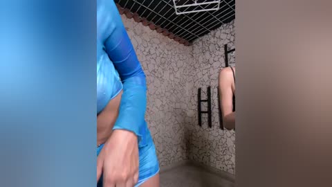 Media: A video of a woman with fair skin in a tight blue athletic top, standing in a tiled shower with a stone-patterned wall. The image is slightly blurred.