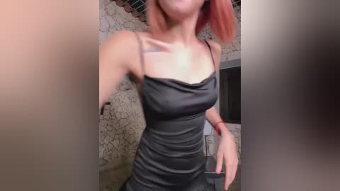 Media: Video of a slender, light-skinned woman with shoulder-length, dyed red hair, wearing a tight, black, spaghetti-strap dress, standing in front of a textured, beige wall with a brick pattern.