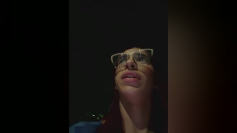 Media: Video of a woman with light skin and brown hair wearing large, reflective glasses, her face slightly tilted upward, against a dark, blurry background.