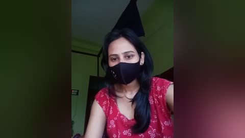 Media: Video of a South Asian woman with long black hair, wearing a black mask, red floral dress, and sitting indoors, with a green wall and dark ceiling in the background.