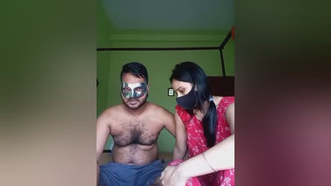 Media: Video of two Indian men, one shirtless with dark hair, wearing a transparent face shield, sitting on a bed. The other, in a red floral dress and face mask, sits beside him. Background features green walls and a wooden chair.