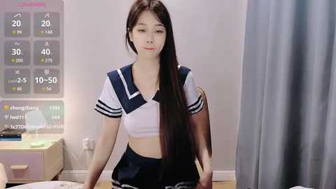 Media: Video of an Asian woman with long, straight black hair, wearing a schoolgirl-style sailor outfit. She sits in a modern, minimalistic room with a white wall and a grey curtain. A digital temperature display is visible in the background.