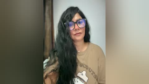 Media: Video of a middle-aged woman with long black hair, wearing glasses, a beige T-shirt, and standing against a beige wall.