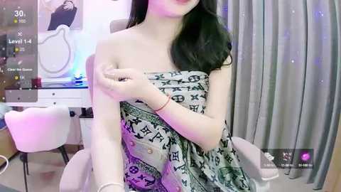 Media: Video of a fair-skinned woman with long black hair, wearing a strapless dress with a pattern, sitting in a modern living room with gray curtains and a vanity table.